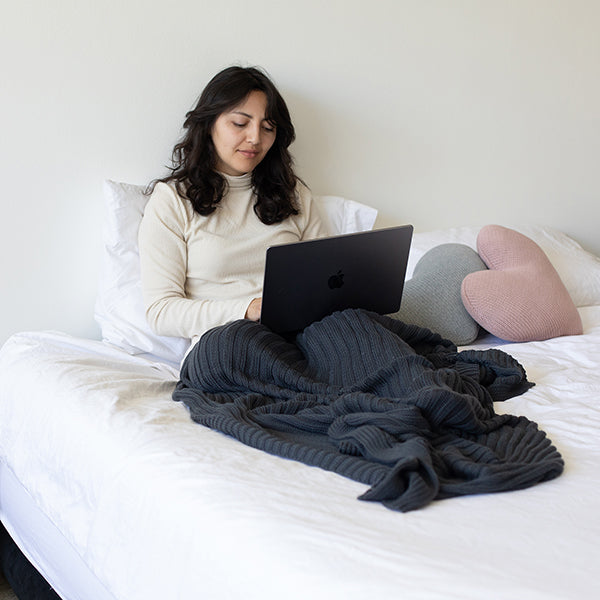 Woman using a laptop on a bed with a blue ribbed throw blanket and heart shaped cushions.