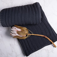 Dark gray and blue knitted cotton blanket with a ribbed texture and a large dried flower on a light marble background.