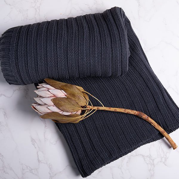 Dark gray and blue knitted cotton blanket with a ribbed texture and a large dried flower on a light marble background.