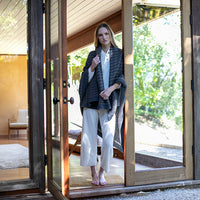 Woman standing in a doorway of a bright house, wearing a white outfit with a dark gray and brown throw blanket.