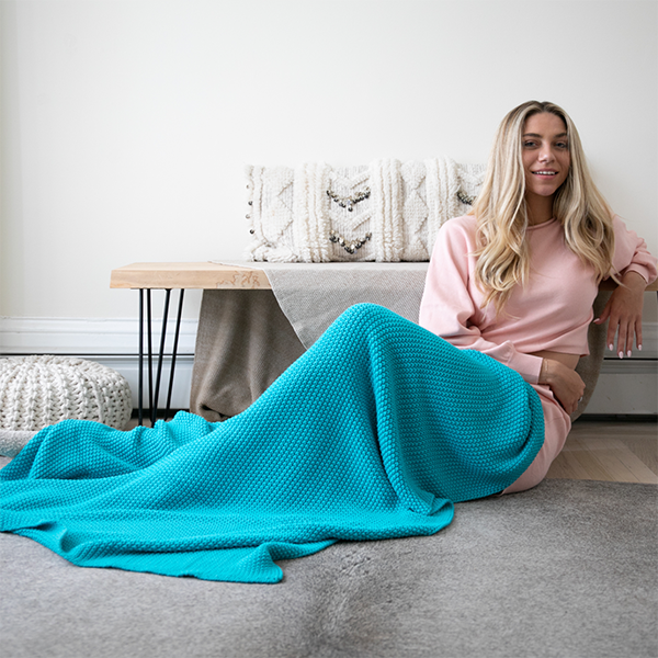 Woman sitting on a couch with a blue blanket draped over her, in a cozy living room setting.