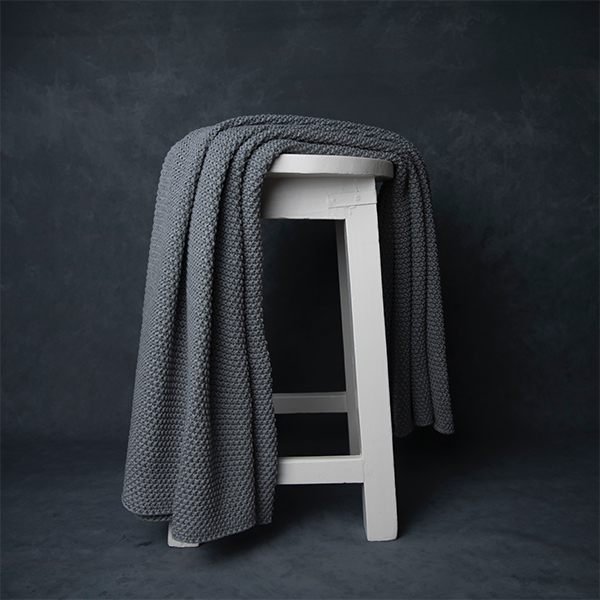 Chunky knitted light grey organic cotton throw blanket draped over a white stool against a dark background.