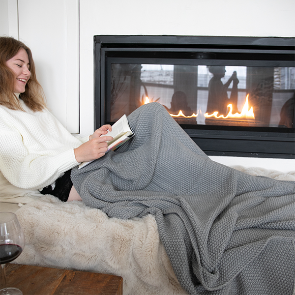 Woman enjoying a book on a couch next to a warm fireplace with a gray chunky knitted throw.