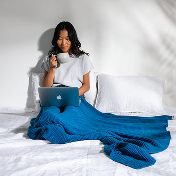 Woman sitting on a bed and sipping on coffee while working on a Apple laptop while wearing a warm marine blue chunky knitted throw.