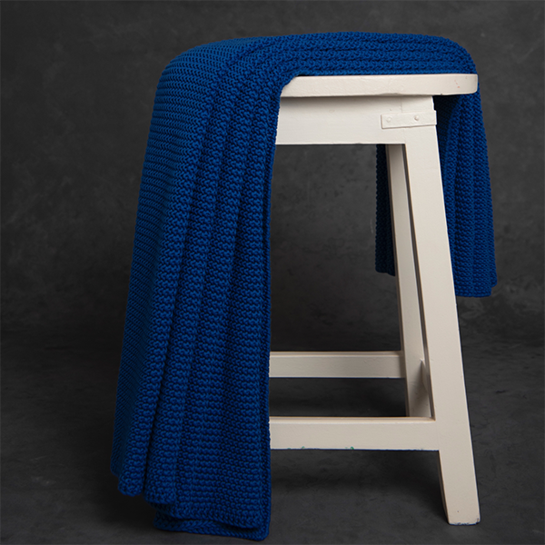 Chunky knitted marine blue organic cotton throw blanket draped over a white stool against a dark background.