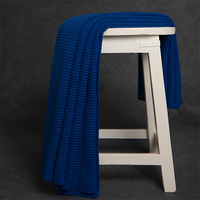 Chunky knitted marine blue organic cotton throw blanket draped over a white stool against a dark background.