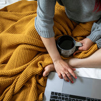 Person using a laptop next to a mustard chunky cotton throw, holding a coffee mug.