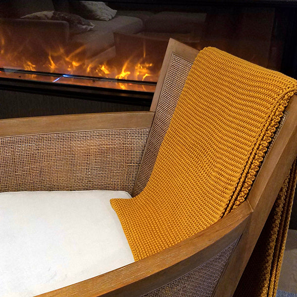 Mustard yellow chunky knitted cotton throw blanket draped over a jute chair in front of a fireplace.