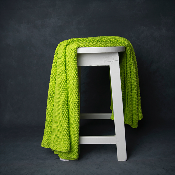 Chunky knitted neon yellow organic cotton throw blanket draped over a white stool against a dark background.