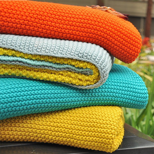Stack of colorful chunky cotton throw in orange, yellow, blue, and white on a surface with a blurred natural background.