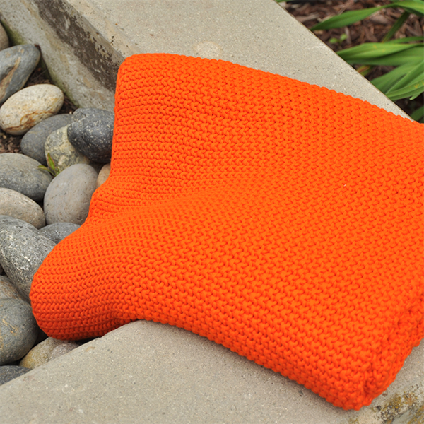 Folded bright orange knitted throw on a stone surface with a natural background next to plants and pebbles.