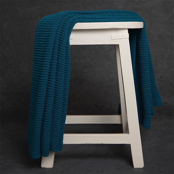Chunky knitted teal petrol blue cotton throw blanket draped over a white stool against a dark background.