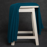 Chunky knitted teal petrol blue cotton throw blanket draped over a white stool against a dark background.