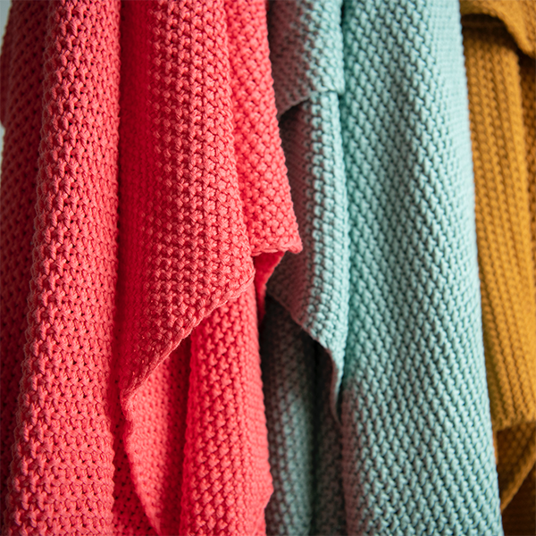 Three chunky knitted throws in red, blue, and mustard pictured next to each other. 