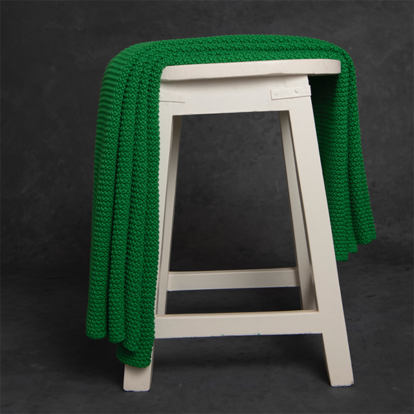Chunky knitted scenic green organic cotton throw blanket draped over a white stool against a dark background.