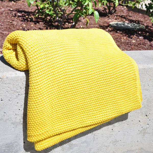 Bright yellow chunky knitted cotton throw on a concrete outdoor surface with plants in the background.