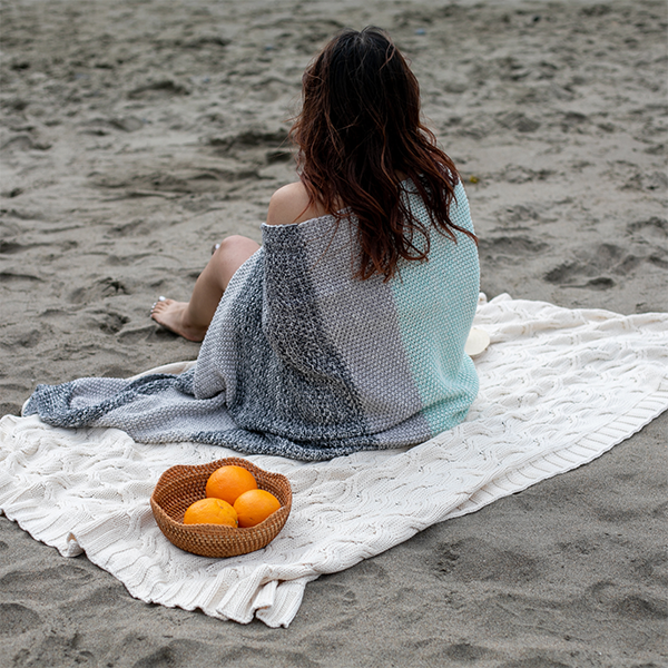 Person sitting on a sandy beach with a mint blue and grey textured blanket and a basket of oranges.
