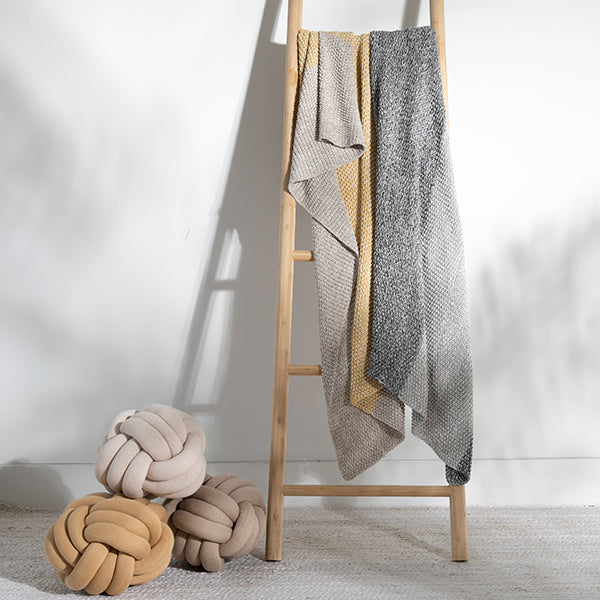 Yellow knit throw blanket on a wooden ladder against a white wall with three pastel coloured knot cushions.