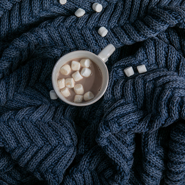 Cup of hot chocolate with marshmallows placed on a chunky blue knitted throw with chevron patterns.