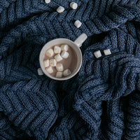 Cup of hot chocolate with marshmallows placed on a chunky blue knitted throw with chevron patterns.