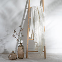 White chunky cotton blanket with chevron patterns draped over a wooden ladder against a white wall with a black vase and white container on the floor.