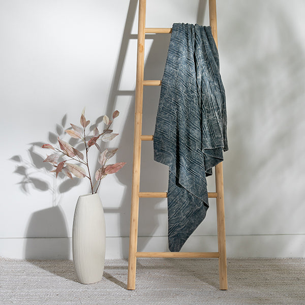 Wooden ladder with a washed blue flintstone throw draped over it next to a white vase with dried plants.