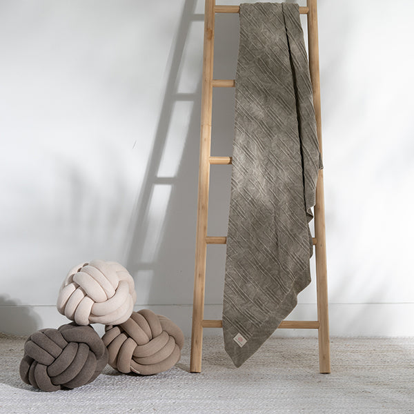 Wooden ladder with a washed cotton stone brown throw draped over it next to a three knot cushions.