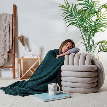 Woman wrapped in a emerald green knit throw laying next to a large grey knot cushion with a plant and books in the background.
