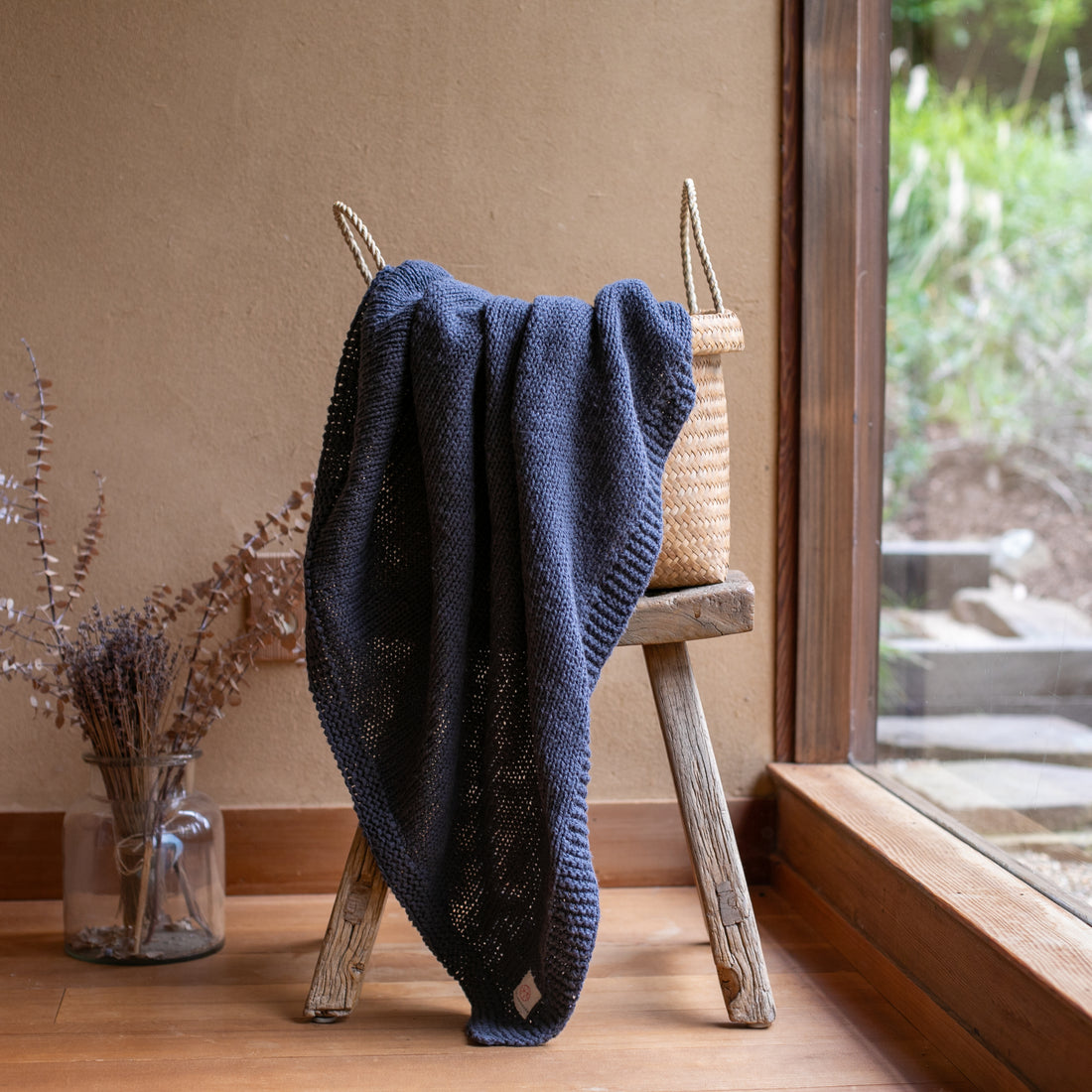 Dark blue Zrestha cotton knit throw hanging out of a jute picnic basket next to a large window. 