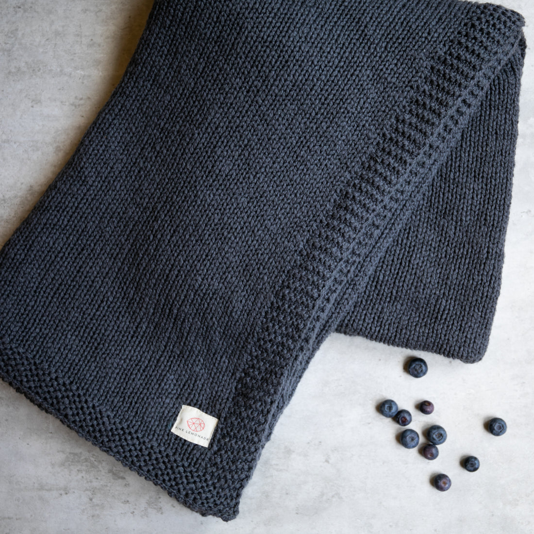 A folded knitted cotton throw blanket in dark blue, with a Pink Lemonade label, displayed on a concrete surface with a few blueberries scattered nearby.
