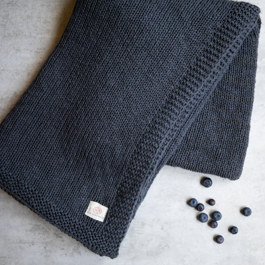 A folded knitted cotton throw blanket in dark blue, with a Pink Lemonade label, displayed on a concrete surface with a few blueberries scattered nearby.