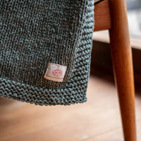 Close up of a dark green knit cotton throw blanket with a Pink Lemonade brand label in the corner.