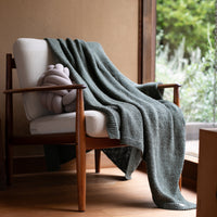 Dark green organic cotton throw blanket placed on top of a wooden chair with a light gray knot cushion.
