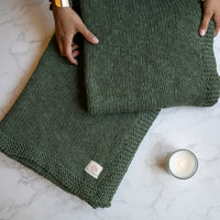 A green knitted cotton throw blanket laid out on a marble surface with a person's hands at the edges and a candle nearby.