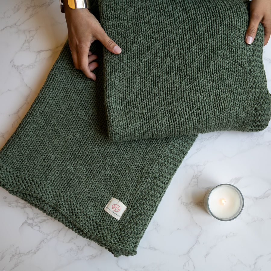A green knitted cotton throw blanket laid out on a marble surface with a person's hands at the edges and a candle nearby.