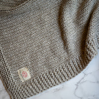 Close up of a Zrestha organic cotton knitted throw in linen grey with a Pink Lemonade brand logo in the corner.