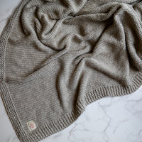 A grey and linen coloured knitted cotton throw blanket draped over a surface, showcasing its textured pattern.