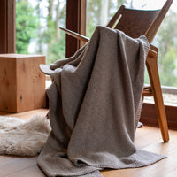 Stone gray blanket draped over a wooden chair in a room with large windows and natural light.