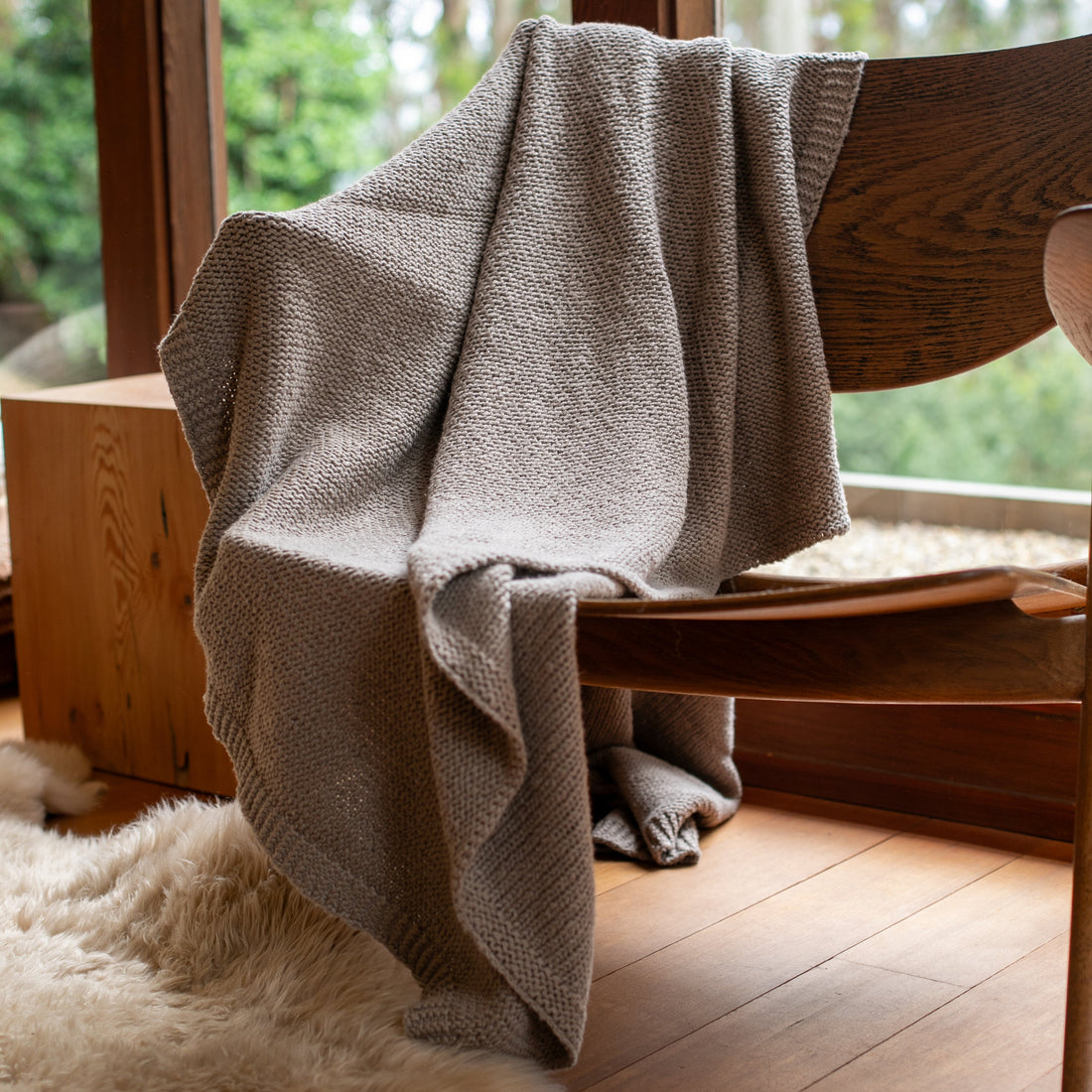 Stone gray cotton throw draped over a wooden chair with a view of trees outside a window.