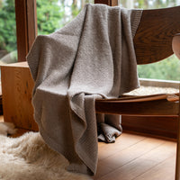 Stone gray cotton throw draped over a wooden chair with a view of trees outside a window.