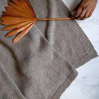 Woven organic cotton throw in stone grey with a dried plant on top on a white marble surface. 