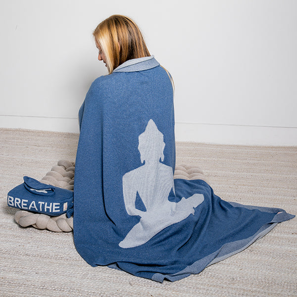 Woman wrapped in a marine blue blanket with a light grey Buddha silhouette, sitting on a knotted mat with a blue pouch saying 'Breathe'.