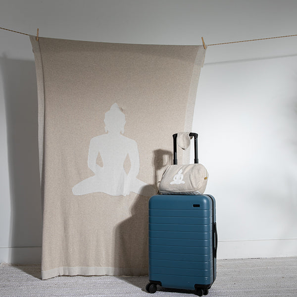 Blue suitcase with a white travel pouch on top, next to a beige linen cotton blanket with a Buddha silhouette.