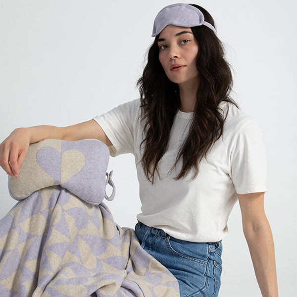 Woman wearing a lavender sleep mask and holding a blanket pouch with a heart and wearing a grey blanket with geometric patterns on a plain background.