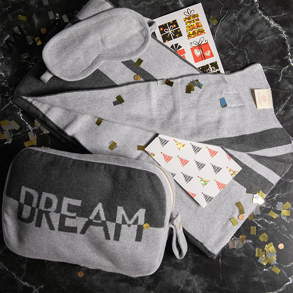 Gray hooded travel blanket, eye mask, and cotton pouch with 'Dream' printed on it on a dark granite surface.