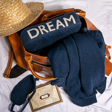 Navy blue travel blanket and pouch with 'DREAM' text, straw hat, brown leather bag and map on a white surface.