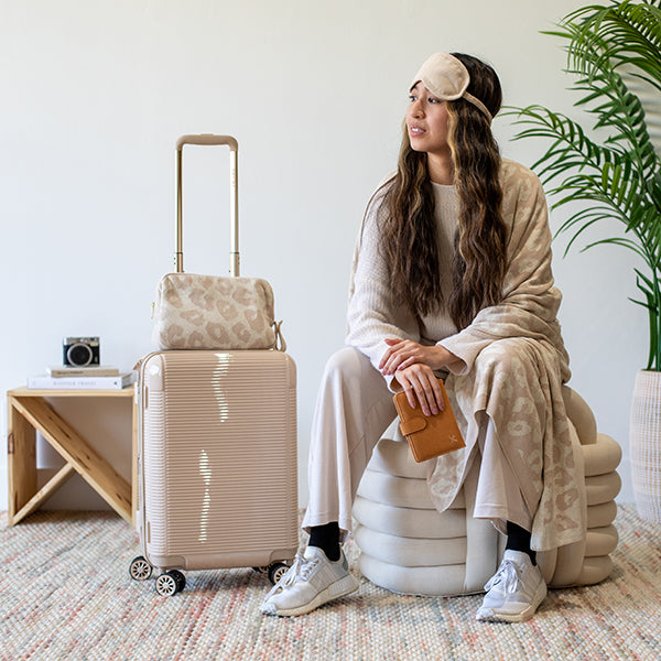 Woman sitting on a beige knot cushion with a suitcase and wearing an eye mask and a leopard print blanket.