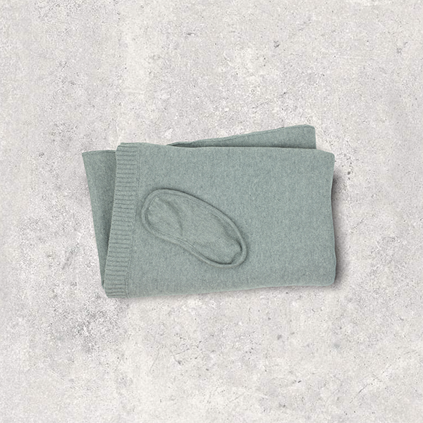 Folded organic cotton light grey travel blanket with matching eye mask on a textured gray surface.
