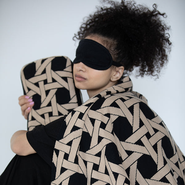 Woman wearing a black eye mask and a patterned black and beige blanket while holding a matching blanket cover against a white background