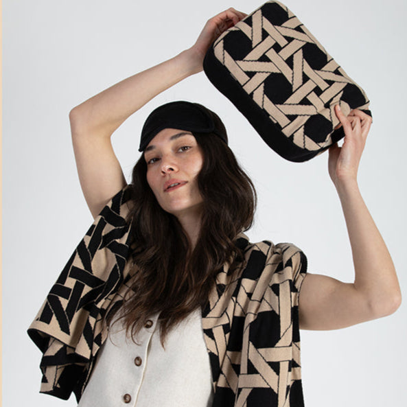 Woman holding a geometric-patterned black travel pouch with a matching cotton blanket wrapped around her.